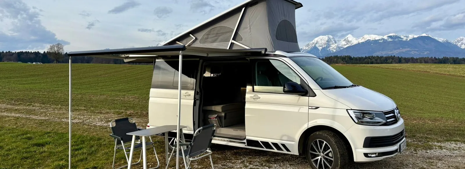VW California Ocean z dvignjeno streho in tendo na travniku pod Kamniškimi Alpami